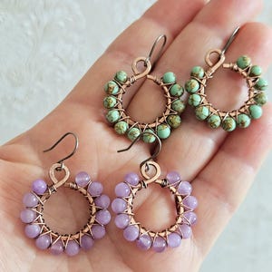 Può includere: Orecchini fatti a mano con un design circolare. Sono visibili due paia di orecchini, uno con perline lavanda e l'altro con perline turchesi. Le perline sono infilate attorno a una montatura in filo di rame.