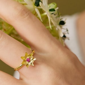 以下が含まれることがあります： 金色のリング。中央に小さな赤い宝石が付いた花のモチーフ。リングは指にあり、緑色の茎とつぼみのある白い花のブーケの隣にあります。