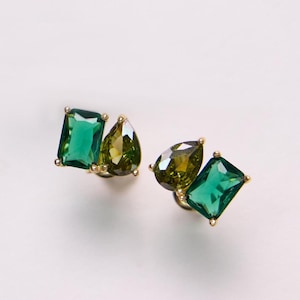 May include: A pair of gold-toned stud earrings, each featuring two gemstones. One earring has a rectangular emerald-green stone and a pear-shaped olive-green stone. The other earring has a rectangular emerald-green stone and a pear-shaped olive-green stone.