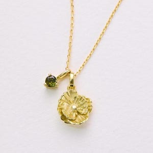 May include: A delicate gold necklace featuring two pendants. One pendant is a small, round, green gemstone, and the other is a detailed, flower-shaped charm. The necklace is displayed on a white surface, with soft, blurred greenery in the background.