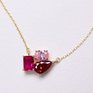 May include: A delicate gold necklace featuring a cluster of colorful gemstones. The necklace showcases a vibrant ruby-red rectangular stone, a light pink oval stone, and a deep red teardrop-shaped stone. The chain is a fine, gold link.