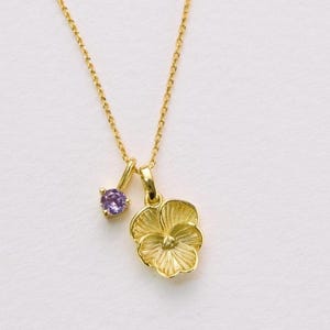 May include: A gold necklace with a flower pendant and a small purple gemstone. The flower pendant has detailed petal textures. The necklace is on a fine chain. The jewellery is set against a white background.