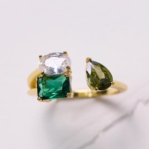 May include: A gold ring with three gemstones: a clear, rectangular stone, a green, rectangular stone, and a green, teardrop-shaped stone. The ring is set against a white background.