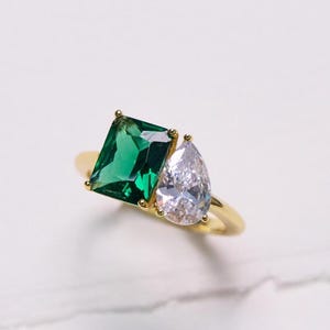 May include: A gold ring featuring a large emerald-cut green gemstone and a smaller pear-shaped clear gemstone. The ring is set on a thin gold band, with the gemstones positioned side-by-side.