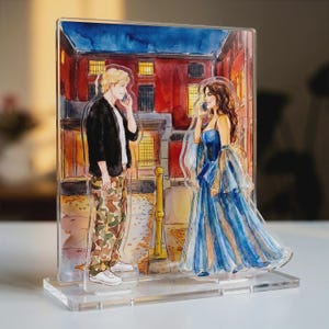 May include: A clear acrylic standee featuring a watercolour illustration of two figures talking on phones. One figure wears a black jacket and camouflage trousers, while the other wears a blue gown. They stand before a red brick building at night.