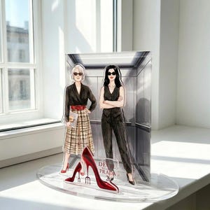 Peut inclure: Présentoir en acrylique avec deux illustrations de mode dans un ascenseur, avec une paire de talons hauts rouges devant. Le texte "The Devil Wears Prada 20" est visible. Les personnages portent des tenues élégantes.