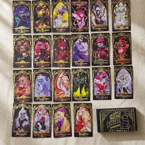 Op de afbeelding: Een verzameling tarotkaarten met kleurrijke illustraties van verschillende personages en symbolen. Elke kaart heeft een uniek ontwerp met gouden accenten en tekst. Een zwarte doos met gouden letters met de tekst "HAPPY HOTEL" is ook inbegrepen.