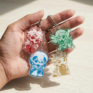 May include: Four acrylic keychains with anime-style characters in red, green, blue, and gold. Each keychain has a silver chain. The characters have distinct hairstyles and outfits. The keychains are held in a hand.