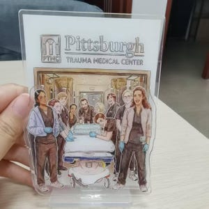 May include: Clear acrylic stand featuring an illustration of a medical team with the text "Pittsburgh Trauma Medical Center". The illustration depicts a group of medical professionals around a hospital bed.