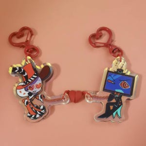 Può includere: Due ciondoli in acrilico collegati da una chiusura magnetica a forma di cuore rosso. Il ciondolo a sinistra raffigura un personaggio con cappello e cappotto arancione e occhi a cuore. Quello a destra mostra uno schermo televisivo blu.