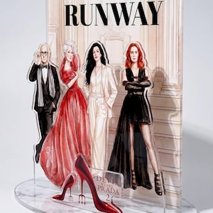 Peut inclure: Présentoir en acrylique transparent avec des illustrations de personnages du film "Le Diable s'habille en Prada" et le mot "RUNWAY" en haut. Le présentoir comprend une paire d'escarpins rouges et une base en marbre.