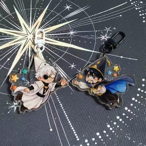 Puede incluir: Dos llaveros acrílicos con dibujos de magos. Un mago viste un atuendo blanco con parche en el ojo, el otro un atuendo azul y negro. Ambos tienen detalles de estrellas y cierres metálicos.