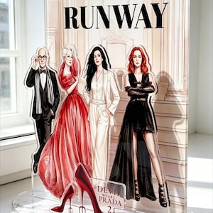 Puede incluir: Expositor acrílico con ilustraciones de personajes de la película "El diablo viste de Prada". El expositor incluye la palabra "RUNWAY" y un par de tacones rojos. También es visible el texto "THE DEVIL WEARS PRADA 2".