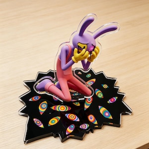 Puede incluir: Una figura de personaje de conejo morado con guantes amarillos, arrodillada sobre una base negra dentada decorada con patrones coloridos en forma de ojos concéntricos. El personaje tiene orejas largas, overol rosa y líquido negro goteando de sus ojos.