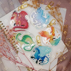 Può includere: Set di cinque portachiavi in acrilico trasparente con creature fantasy: un drago rosso, un unicorno blu, un serpente marino verde, una fenice arancione e un grifone blu. Ogni portachiavi ha un fermaglio dorato ed è decorato con glitter.
