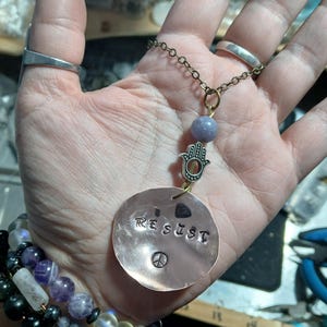 May include: A copper pendant necklace with the word "RESIST" stamped on it, along with a peace symbol. The necklace features a small Hamsa hand charm, a purple bead, and a bronze chain. A beaded bracelet with various colored beads is also visible.