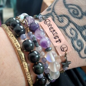 May include: A collection of beaded bracelets in shades of black, purple, and white. One bracelet features a copper tag with the word "RESIST" and a peace sign. Another bracelet is a gold-colored cuff with engraved text.