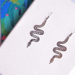 May include: A pair of silver snake earrings with a delicate floral design. The earrings are dangle style and have a simple hook fastening.