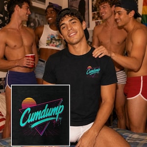 May include: A black short-sleeved t-shirt featuring a graphic print on the chest. The design displays a stylised sunset above a purple triangle with a grid pattern and the text "Cumdump" in a bright teal, retro-inspired cursive font.