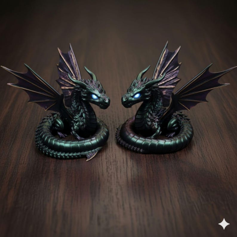 Pu&ograve; includere: Due dettagliate statuette di drago. Ogni drago &egrave; verde scuro con ali nere e occhi blu luminosi. I draghi sono arrotolati e si trovano su una superficie di legno scuro. Le ali sono spiegate e i draghi si guardano.