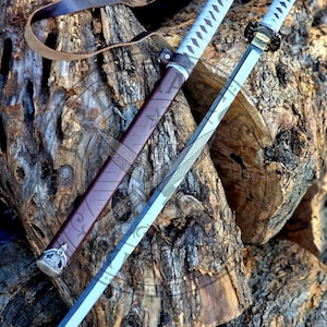 Puede incluir: Dos espadas katana con empuñaduras azul claro y hojas plateadas sobre una superficie de madera envejecida. Las vainas son marrones con detalles plateados. Se ve una correa de cuero. Estas espadas son probablemente para exhibición o artes marciales.