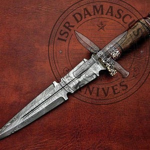 Puede incluir: Una daga de doble filo con una hoja de acero damasco estampada que presenta un canal central. El mango está hecho de madera oscura con virolas metálicas intrincadas y un pomo. El texto ISR DAMASCUS KNIVES es visible en el fondo.