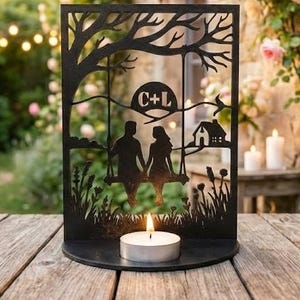 May include: Black metal candle holder featuring a silhouette of a couple on a swing under a tree, with initials "C+L" inside a circle. A small house and a lit candle are also visible. The holder sits on a wooden table in a garden setting.