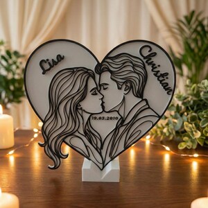 Puede incluir: Decoraci&oacute;n de madera en forma de coraz&oacute;n con un dise&ntilde;o en capas que representa a una pareja bes&aacute;ndose. Los nombres "Lisa" y "Christian" est&aacute;n inscritos, junto con la fecha "19.03.2010". El dise&ntilde;o en blanco y negro est&aacute; montado sobre una base blanca.