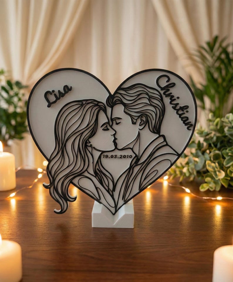Puede incluir: Decoraci&oacute;n de madera en forma de coraz&oacute;n con un dise&ntilde;o en capas que representa a una pareja bes&aacute;ndose. Los nombres "Lisa" y "Christian" est&aacute;n inscritos, junto con la fecha "19.03.2010". El dise&ntilde;o en blanco y negro est&aacute; montado sobre una base blanca.