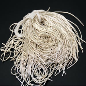 Pode incluir: Um feixe de cordão prateado, enrolado e agrupado com um fio branco. Os cordões são finos e flexíveis, com um brilho metálico. O fundo é preto sólido, realçando a textura e a cor dos cordões.