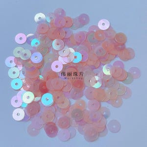 Puede incluir: Una colección de pequeñas lentejuelas redondas de color melocotón con un acabado iridiscente, cada una con un orificio central para coser. El texto 伟丽珠片 Workshop está impreso en el centro del montón de adornos artesanales planos y circulares.