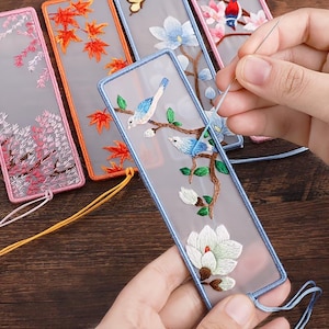 Peut inclure: Marque-pages brodés avec des motifs floraux et d'oiseaux. Chaque marque-page présente un motif différent, notamment des fleurs de cerisier roses, des feuilles d'érable oranges, des oiseaux bleus sur une branche et des magnolias blancs. Les marque-pages ont un fond transparent et des bordures colorées.