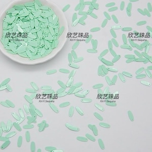Peut inclure: Séquins ovales vert menthe dispersés sur une surface blanche, certains dans un bol blanc. L'image comprend le texte "XinYi Sequins" en anglais et en caractères chinois. Ces sequins sont parfaits pour les projets d'artisanat.