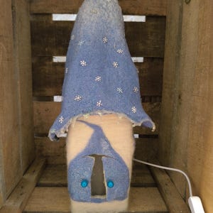 Könnte beinhalten: Handgefertigte Lampe aus blauem und weißem Filz. Die Lampe hat eine spitz zulaufende Hutform, die mit Schneeflocken verziert ist, und ein passendes Gesicht mit blauen Edelsteinaugen. Die Lampe ist mit einem weißen Kabel angeschlossen.