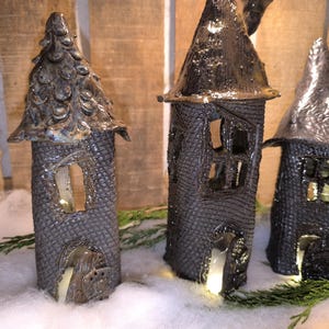 Könnte beinhalten: Drei Keramik-Feenhäuser mit strukturierten, dunkelgrauen Außenwänden und beleuchteten Fenstern und Türen. Die Häuser haben unterschiedliche Dachdesigns, darunter eine konische und eine strukturierte, baumartige Form. Sie befinden sich in einer verschneiten Szene.