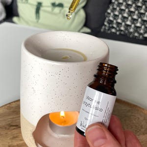 May include: A white and beige essential oil diffuser with a lit candle inside. A small brown bottle of oil and a dropper are held above the diffuser. The bottle has a white label with text.