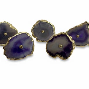 May include: Five decorative cabinet knobs with a natural agate design. The knobs feature a gold-colored edge and a small, gold-colored center post. The agate has shades of purple, gray, and white.