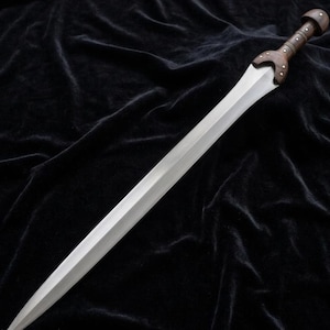 Puede incluir: Una espada larga y plateada con empuñadura y guarda de madera. La hoja es recta y se estrecha hasta un punto. El mango es de color marrón oscuro con remaches. La espada se muestra sobre un fondo negro.
