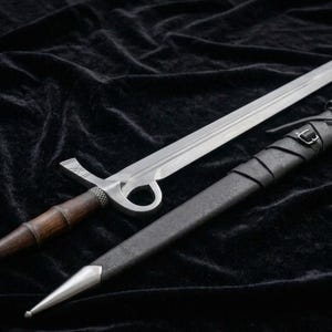May include: A medieval-style sword with a silver blade and a wooden handle. The sword is accompanied by a black leather sheath. The handle has a silver pommel and a guard with a circular design. The sheath is wrapped with leather straps.