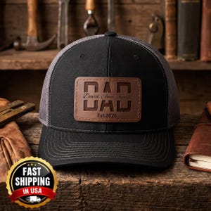 May include: A black baseball cap with a grey mesh back and a rectangular brown leather patch on the front. The patch features the word DAD in large block letters, the names David, Anna, and Kamila in script, and the text Est 2026.