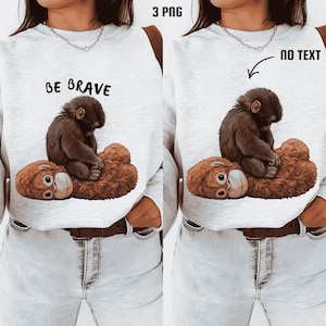 Könnte beinhalten: Hellgraues Sweatshirt mit einem braunen Affen und einem Teddybären-Design. Der Text "BE BRAVE" ist in Schwarz über dem Affen aufgedruckt. Das Sweatshirt hat einen Rundhalsausschnitt und lange Ärmel.