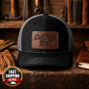 Pode incluir: Um boné de beisebol preto com a parte traseira em rede cinzenta e um emblema retangular de couro castanho na frente. O emblema tem gravadas as palavras CUSTOM TEXT numa fonte escura, a negrito e em estilo cursivo. O boné tem aba curva.