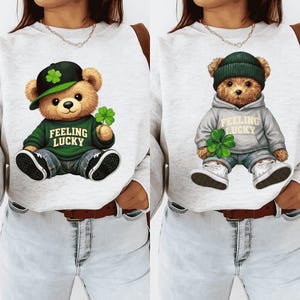 May include: Two light gray sweatshirts with a St. Patrick's Day theme. Each sweatshirt features a cartoon teddy bear wearing a hat and holding a four-leaf clover. The text "Feeling Lucky" is printed on the bear's shirt.