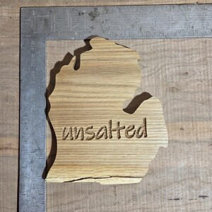 Op de afbeelding: Een houten snijwerk in de vorm van de staat Michigan, met het woord "unsalted" in het hout gesneden. Het hout heeft een natuurlijke nerf en is lichtbruin van kleur. Een metalen liniaal staat op de achtergrond.