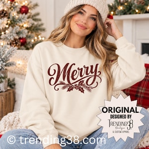 Könnte beinhalten: Ein cremefarbenes Sweatshirt mit Rundhalsausschnitt und dem Wort Merry in einer dunkelroten, abgenutzten Schreibschrift, verziert mit einer Grafik aus Stechpalmenblättern und Tannennadeln. Ein sternförmiges Abzeichen zeigt ORIGINAL DESIGNED BY TRENDING38.