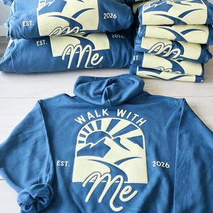 K&ouml;nnte beinhalten: Ein blauer Kapuzenpullover mit einer hellgelben Grafik auf der R&uuml;ckseite, die eine Berglandschaft, eine Sonne und den Text "WALK WITH Me EST. 2026" zeigt. Der &Auml;rmel tr&auml;gt den Text "Moses 6:34". Mehrere gefaltete blaue Pullover sind gestapelt.