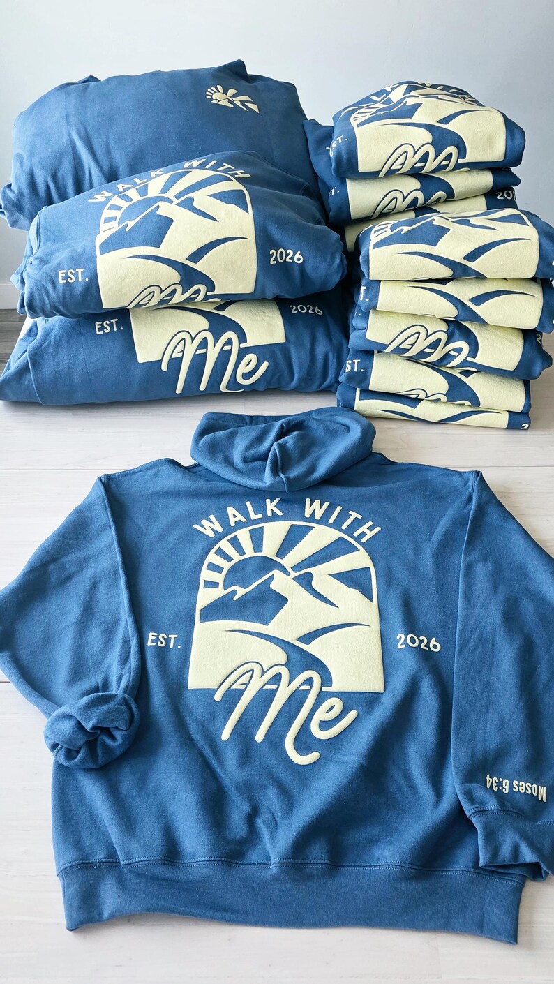K&ouml;nnte beinhalten: Ein blauer Kapuzenpullover mit einer hellgelben Grafik auf der R&uuml;ckseite, die eine Berglandschaft, eine Sonne und den Text "WALK WITH Me EST. 2026" zeigt. Der &Auml;rmel tr&auml;gt den Text "Moses 6:34". Mehrere gefaltete blaue Pullover sind gestapelt.