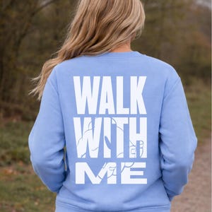 Puede incluir: Sudadera azul claro con la frase "WALK WITH ME" impresa en letras blancas grandes en la espalda. La sudadera es de color liso y tiene cuello redondo.