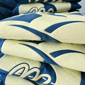 K&ouml;nnte beinhalten: Ein Stapel gefalteter dunkelblauer Sweatshirts mit einem gro&szlig;en, erhabenen, hellgelben Grafikdruck. Das Design besteht aus sich wiederholenden, geschwungenen, abstrakten Formen, die ein kr&auml;ftiges, strukturiertes Muster auf dem Stoff bilden.