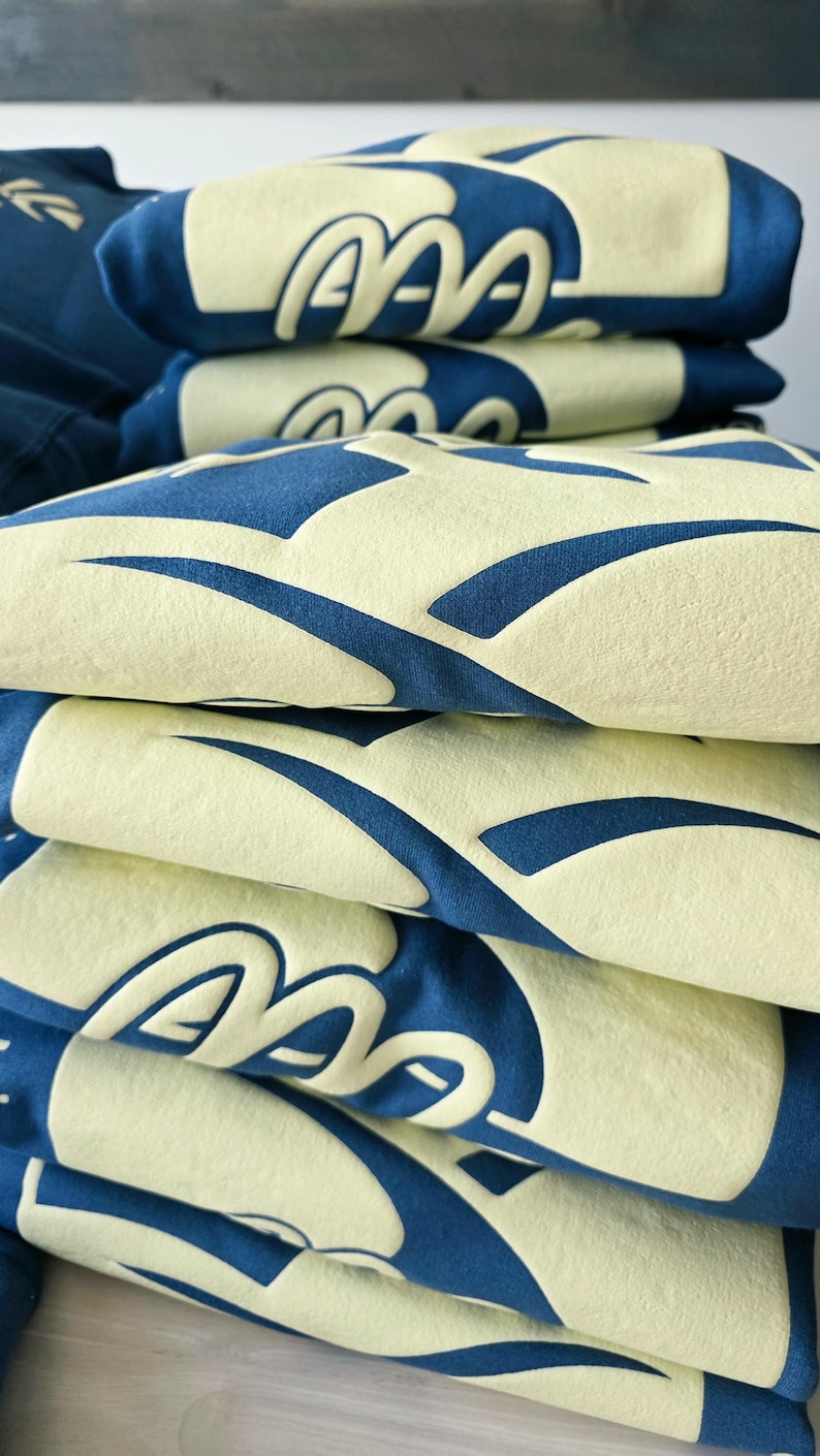 K&ouml;nnte beinhalten: Ein Stapel gefalteter dunkelblauer Sweatshirts mit einem gro&szlig;en, erhabenen, hellgelben Grafikdruck. Das Design besteht aus sich wiederholenden, geschwungenen, abstrakten Formen, die ein kr&auml;ftiges, strukturiertes Muster auf dem Stoff bilden.